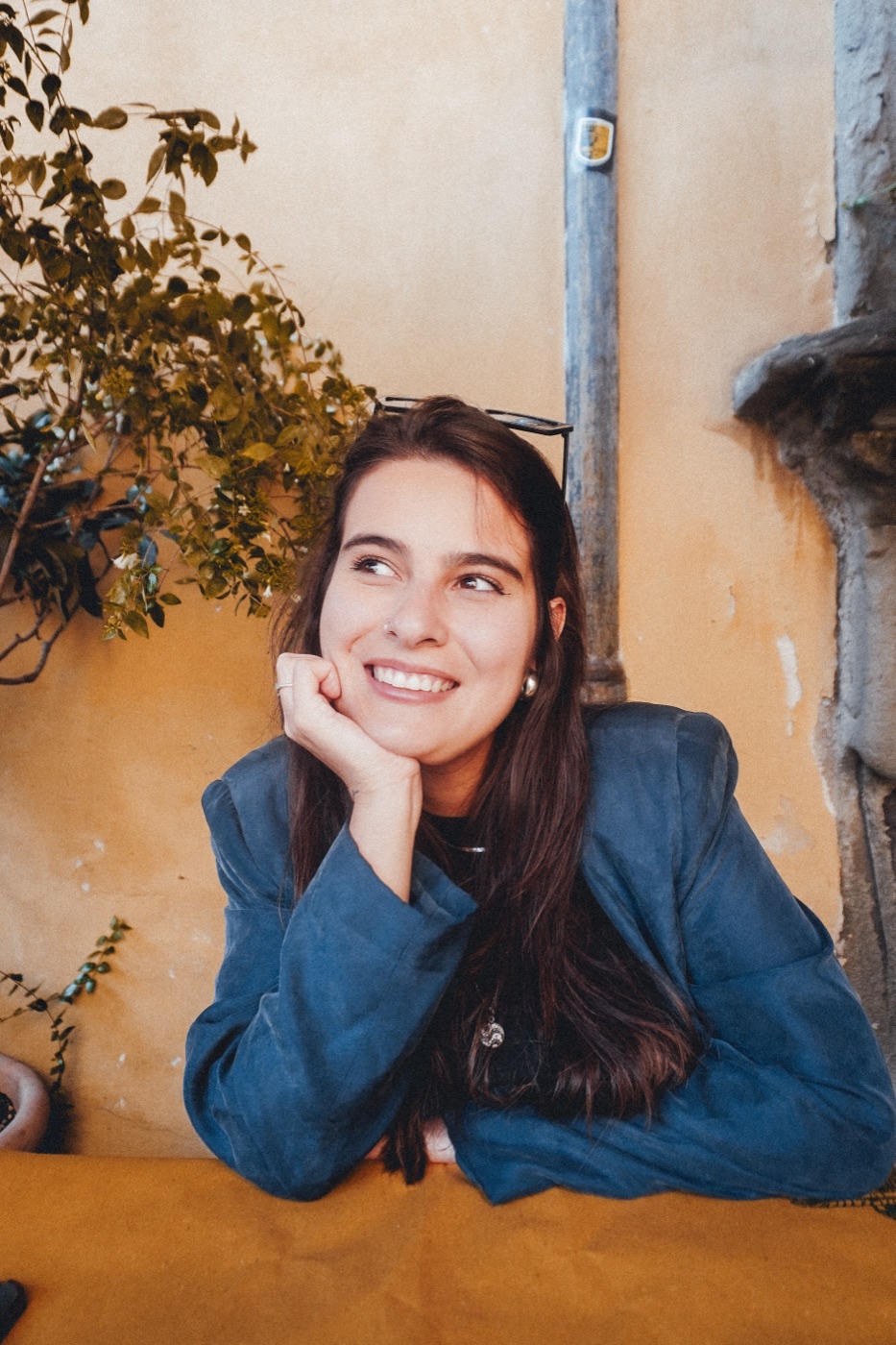 Anna sorrindo, com a mão no queixo, diante de um muro amarelo